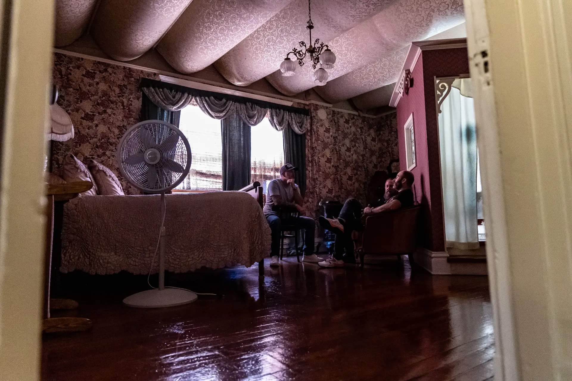 The three hosts sitting in a dimly lit Victorian bedroom of a haunted house