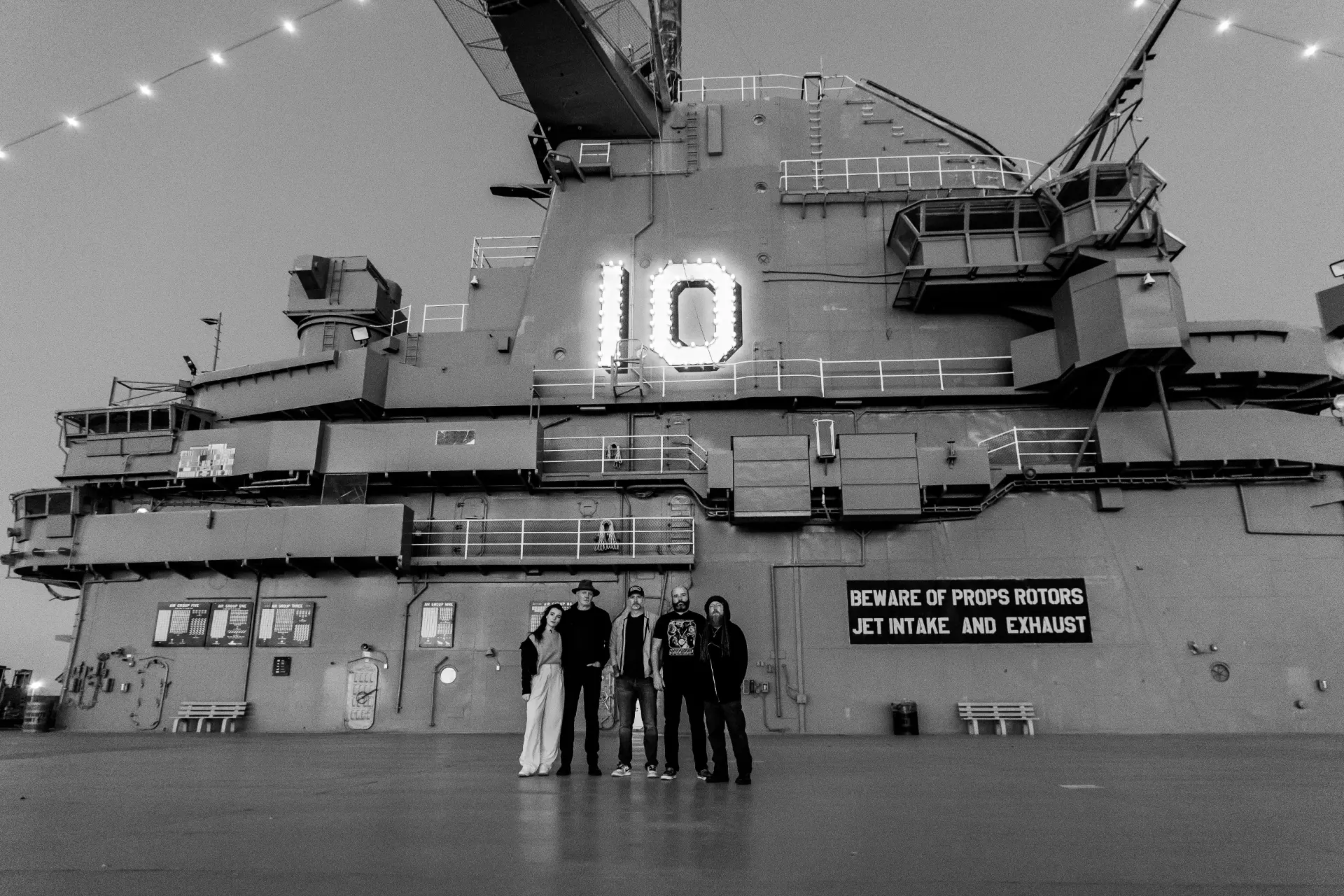 The cast standing on the flight deck of a historic aircraft carrier at dusk